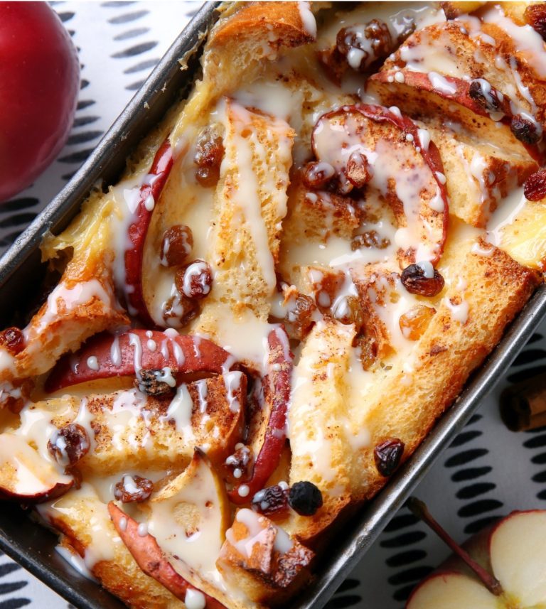Bread pudding with apple slices, raisins, and a white drizzle in a baking dish.